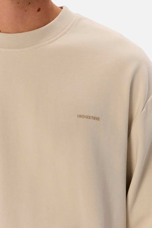 Close-up of a person wearing the beige Industrie The Newport Long Sleeve Tee. The focus is on the chest area, featuring a small printed logo that reads "INDUSTRIE." The fabric appears smooth and soft with a heavy weight finish. The background is plain white.