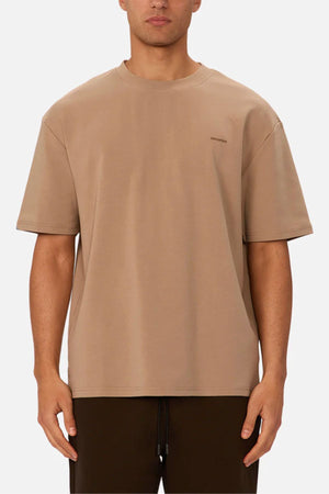 A person wears the Industrie The Newport Tee in beige, featuring an oversized fit, paired with dark pants and standing with arms relaxed by their sides against a plain white background.