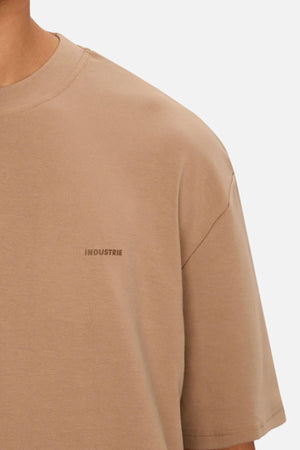 A close-up of someone wearing the Industrie The Newport Tee, an oversized beige 100% cotton t-shirt with a subtle "INDUSTRIE" logo on the chest, set against a plain, light background.