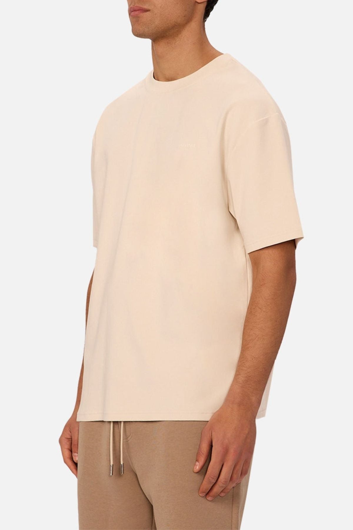 A man, partially out of frame, stands against a plain white background wearing the Industrie The Newport Tee in an oversized fit with beige light brown drawstring pants.