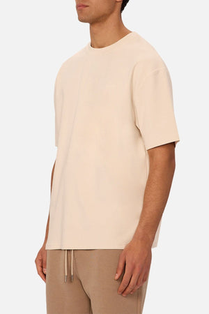 A man, partially out of frame, stands against a plain white background wearing the Industrie The Newport Tee in an oversized fit with beige light brown drawstring pants.