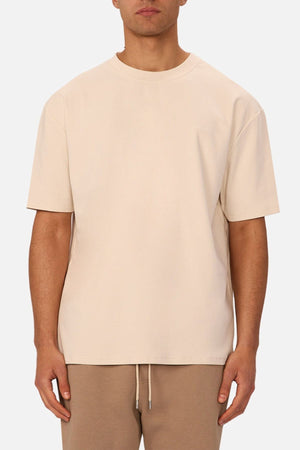A man faces forward wearing an oversized fit, light beige Industrie The Newport Tee and tan drawstring pants against a plain white background.