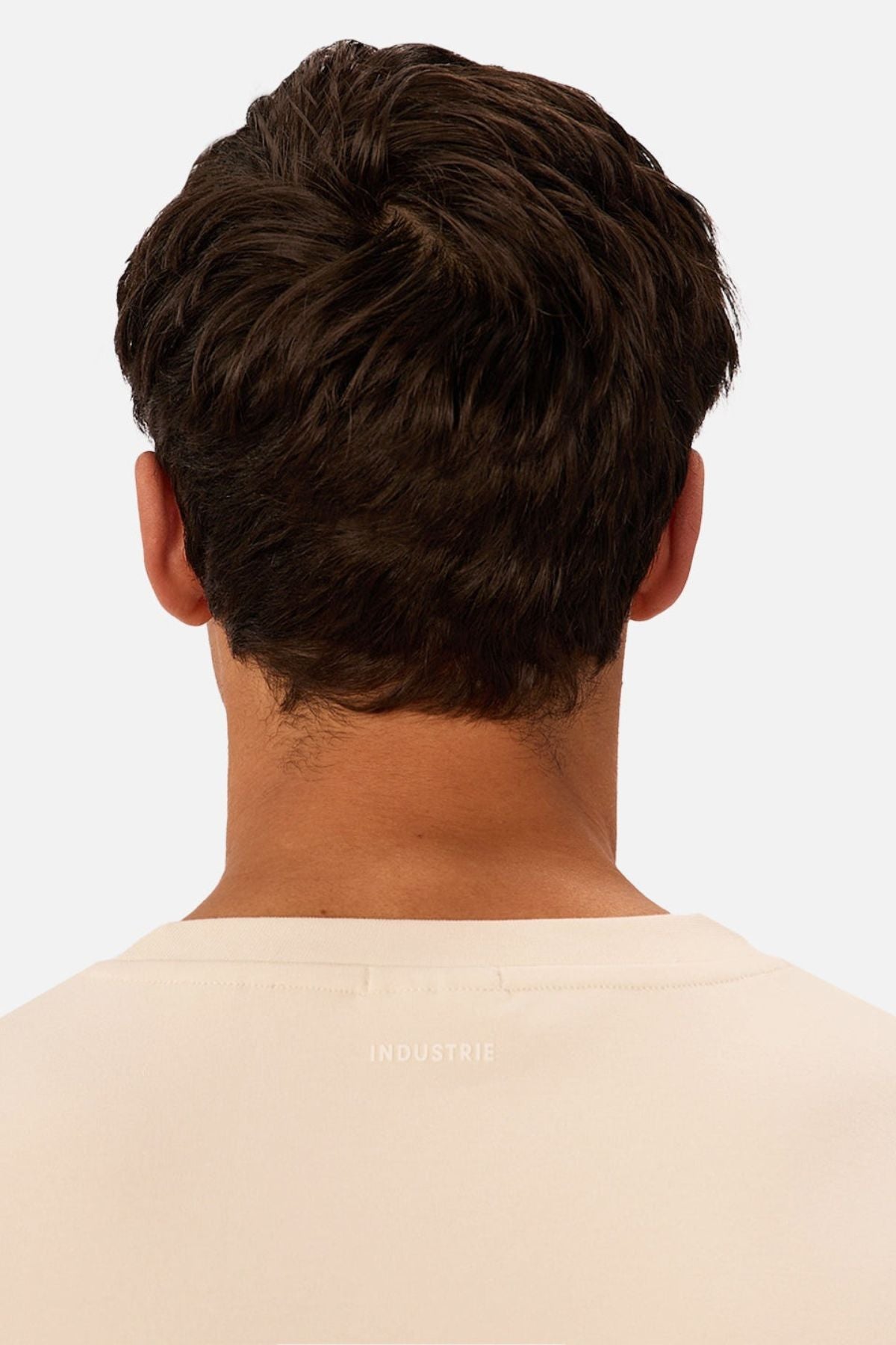 Seen from the back, a person with short dark brown hair wears a cream Industrie The Newport Tee with "INDUSTRIE" near the neckline. The tee has an oversized fit and is set against a plain, light background.
