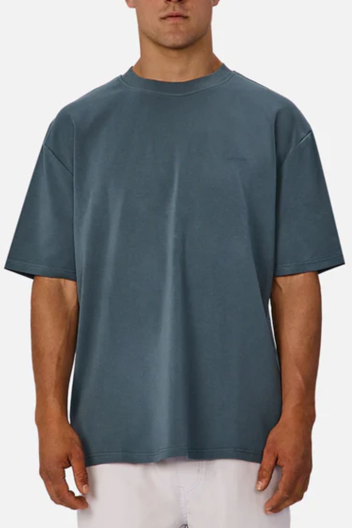 A man wears an oversized Industrie The Newport Tee in blue with light-colored pants, standing against a white background. The tee has a minimal, graphic-free design.