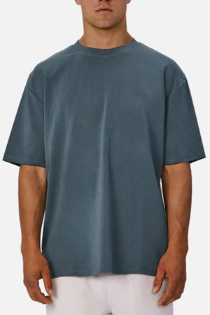 A man wears an oversized Industrie The Newport Tee in blue with light-colored pants, standing against a white background. The tee has a minimal, graphic-free design.