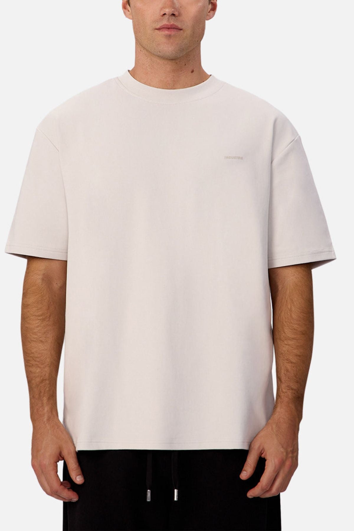A man in an Industrie The Newport Tee, a loose-fitting light beige short-sleeve shirt with subtle chest text and minimalistic design, pairs it with black pants while standing against a plain white background.