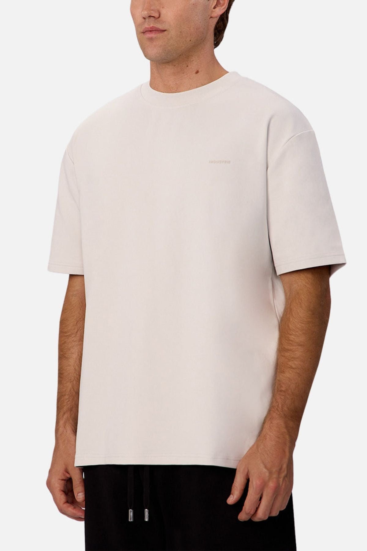 A man wears the Industrie The Newport Tee in light beige, featuring subtle chest text, paired with black pants, standing against a plain white background.