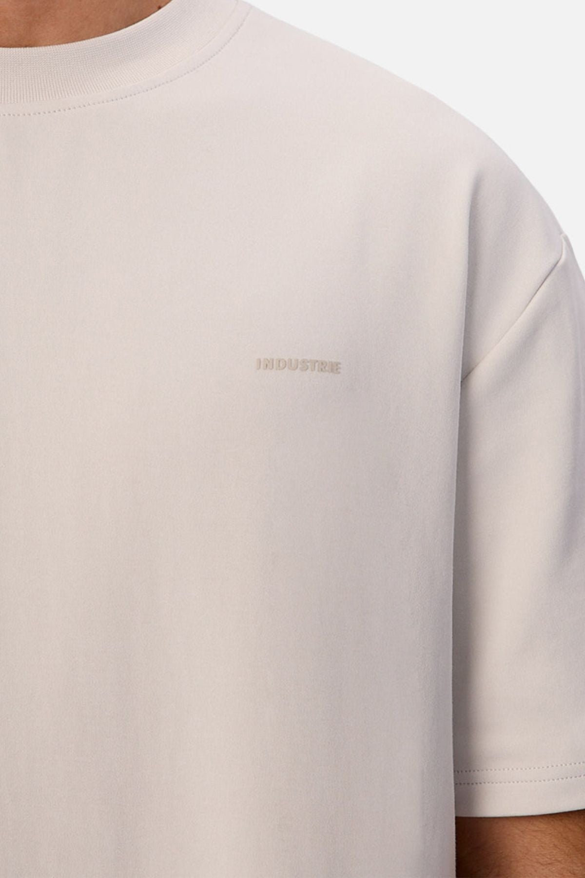 Close-up of a person wearing the Industrie The Newport Tee in light beige, featuring short sleeves, an oversized fit, and "INDUSTRIE" subtly embroidered on the chest. Only their upper body and neck are visible.