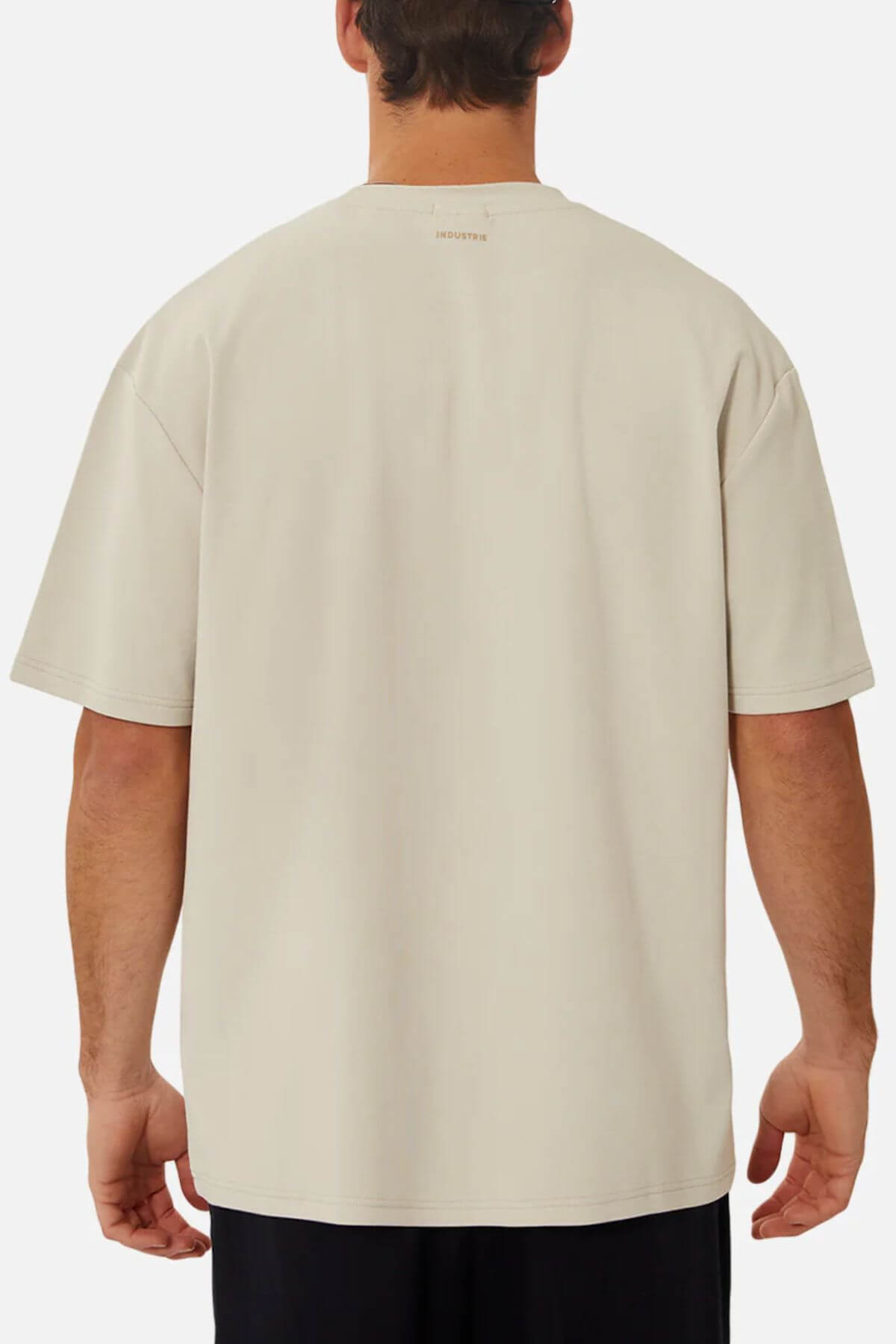 A person is standing with their back facing the camera, wearing an oversized fit, beige Industrie The Newport Tee and dark pants. The 100% cotton T-shirt features a small, barely visible text at the upper back near the neckline. The background is plain and light gray.