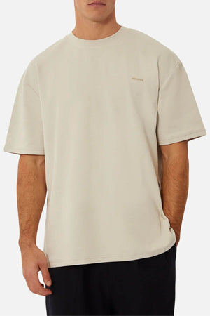 A person is wearing a loose-fitting, 100% cotton beige T-shirt with short sleeves and black pants. The Industrie The Newport Tee features minimal text on the upper right side. The person has their left hand in their pocket and is standing against a plain light background.