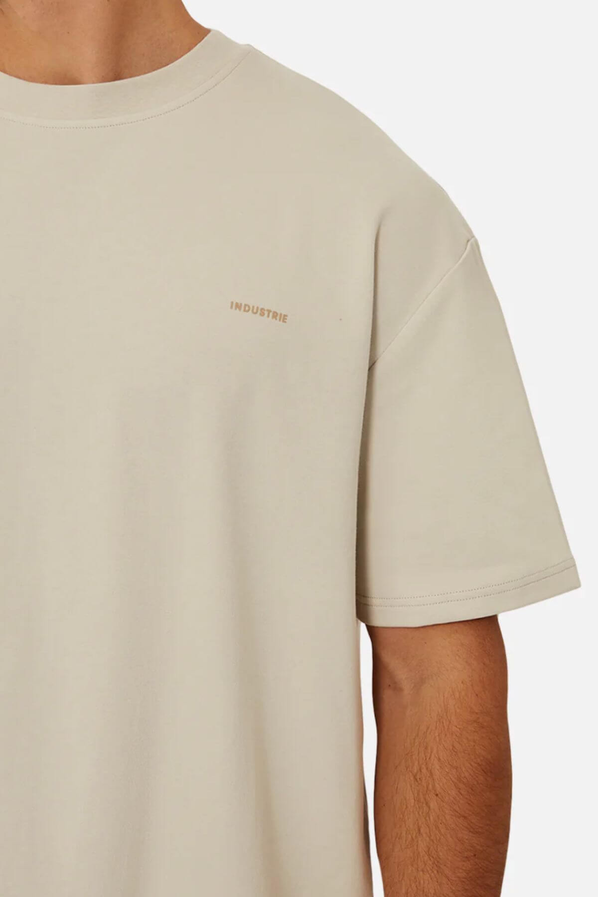 A close-up of a person wearing Industrie The Newport Tee, a light beige t-shirt made from 100% cotton with the word "INDUSTRIE" subtly printed on the left side of the chest. Only the upper torso and part of one arm are visible against a plain background.