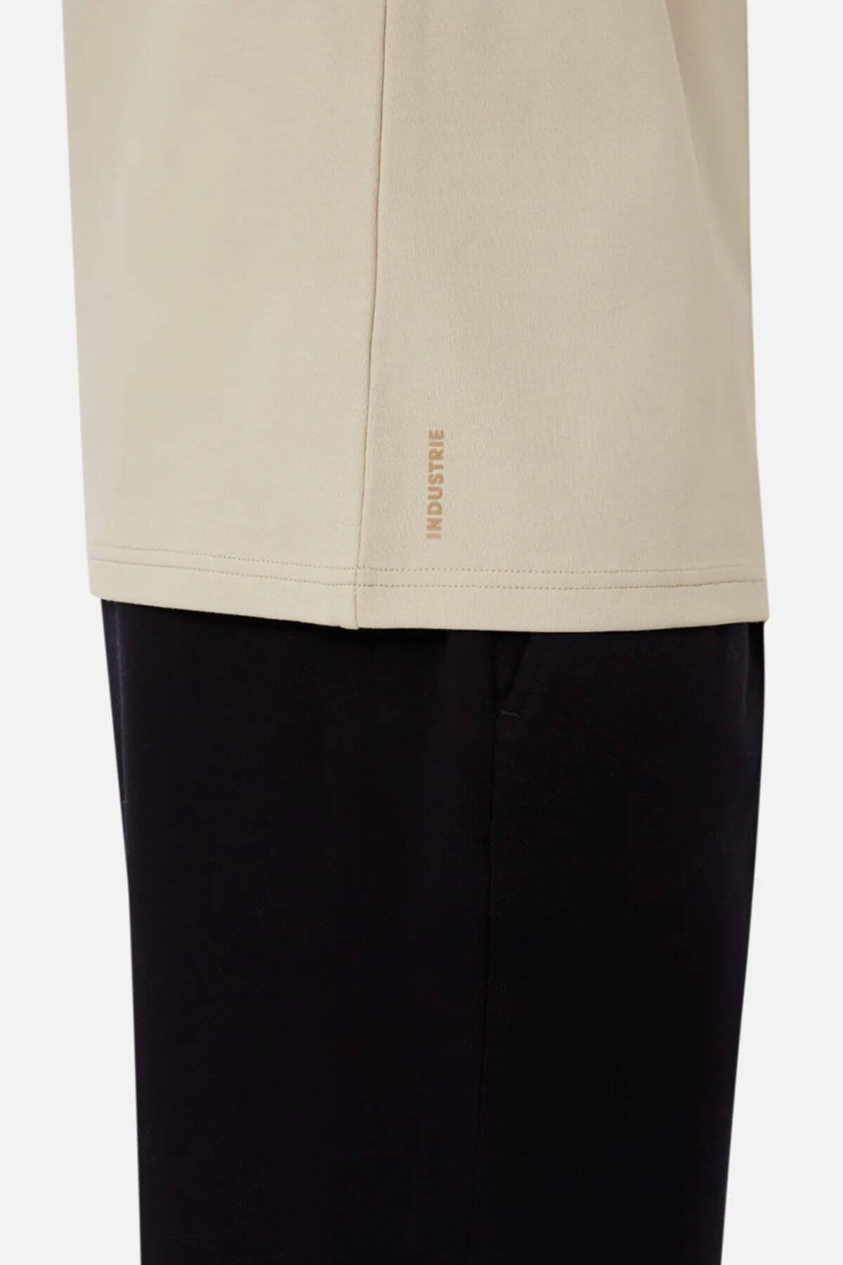 A close-up shot features a person in an oversized fit beige Industrie The Newport Tee paired with black pants. Made from 100% cotton, the shirt displays the "INDUSTRIE" brand name in small letters near the bottom hem on the left side. The image captures only the lower portion of the shirt and the upper section of the pants.