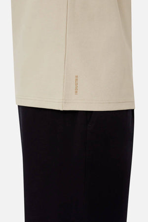 A close-up shot features a person in an oversized fit beige Industrie The Newport Tee paired with black pants. Made from 100% cotton, the shirt displays the "INDUSTRIE" brand name in small letters near the bottom hem on the left side. The image captures only the lower portion of the shirt and the upper section of the pants.