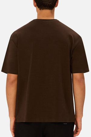 A person is seen from behind wearing an oversized Industrie The Newport Tee in plain dark brown with black pants, set against a light gray background.