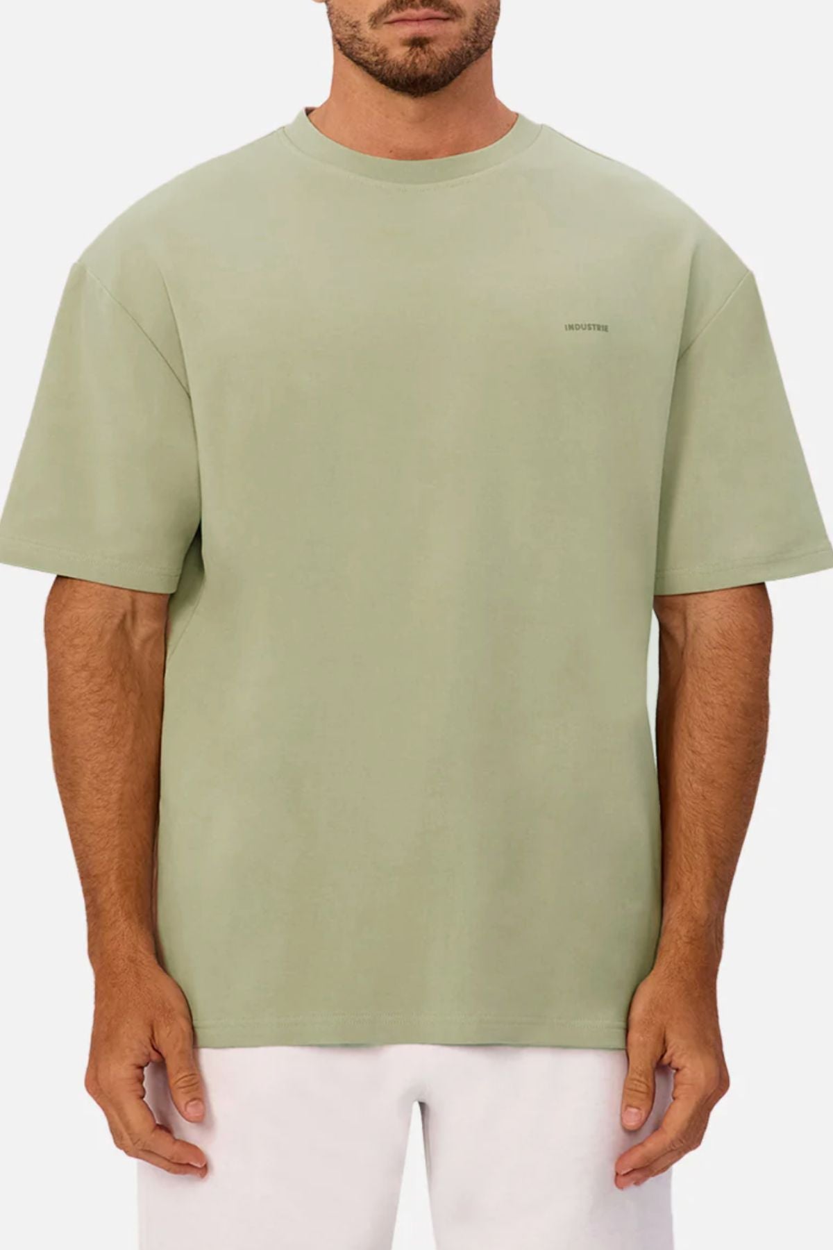 A man faces forward, wearing Industrie The Newport Tee—an oversized sage green heavyweight 100% cotton t-shirt with a subtle left chest logo. He pairs it with white pants, standing against a plain white background.
