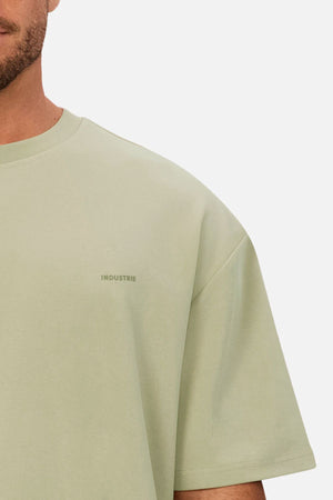 A man wears an oversized Industrie The Newport Tee in light green, 100% cotton, with a subtle "INDUSTRIE" logo on the chest. Only the left side of his upper body and part of his face are visible.