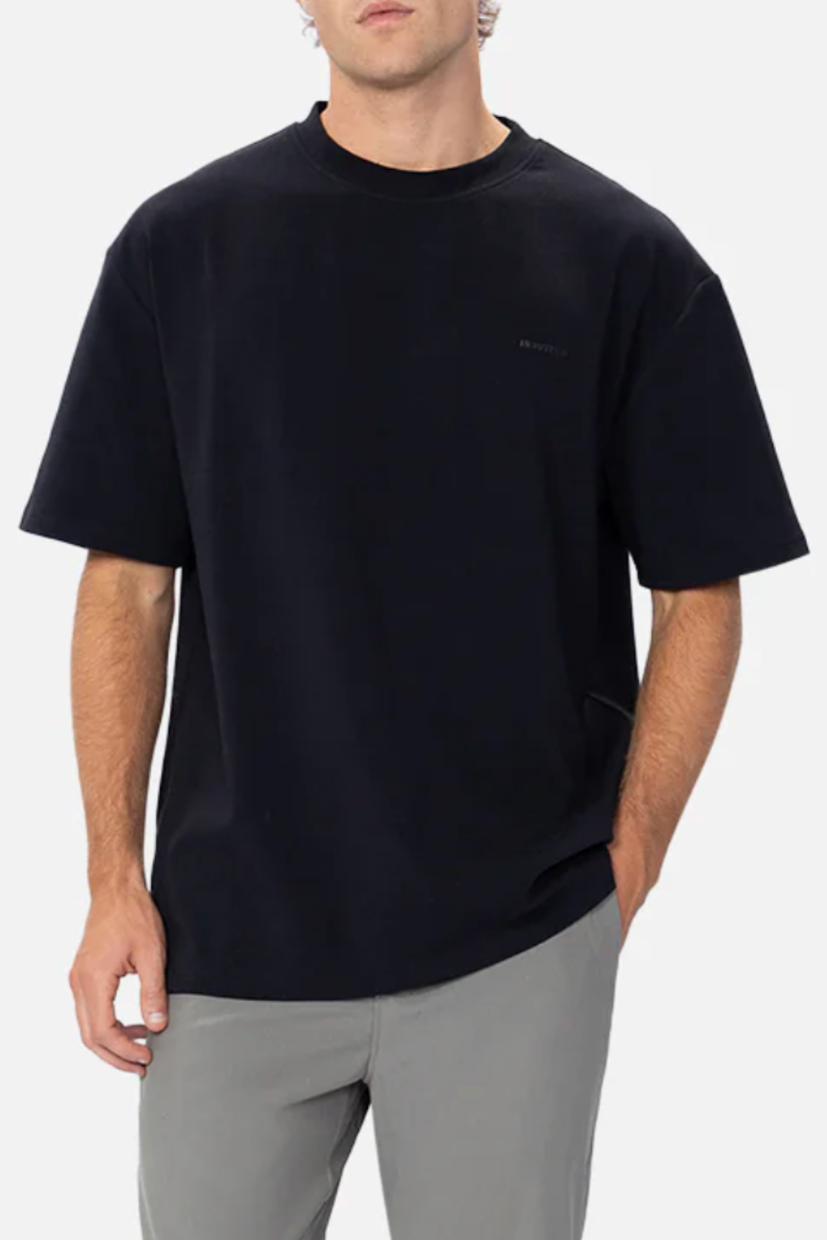 A person with light skin and short hair stands against a plain white background, wearing Industrie The Newport Tee in a loose-fitting, black 100% cotton fabric paired with light gray pants. They have one hand tucked into their pocket and maintain a relaxed posture, with only the upper half of their body visible.