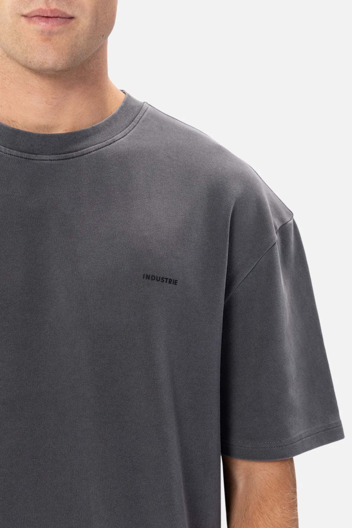 The person wears Industrie The Newport Tee, an oversized 100% cotton T-shirt with "INDUSTRIE" on the chest. Set against a plain white backdrop, it accentuates their upper body and face, highlighting the shirt's sleek, modern design.