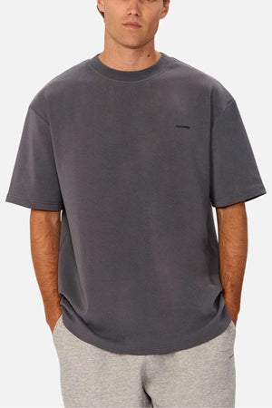 A person stands with their hands in their pockets, wearing an oversized fit, dark gray T-shirt and light gray shorts. The 100% cotton Industrie The Newport Tee features a small, subtle logo on the left side of the chest. The background is plain white.