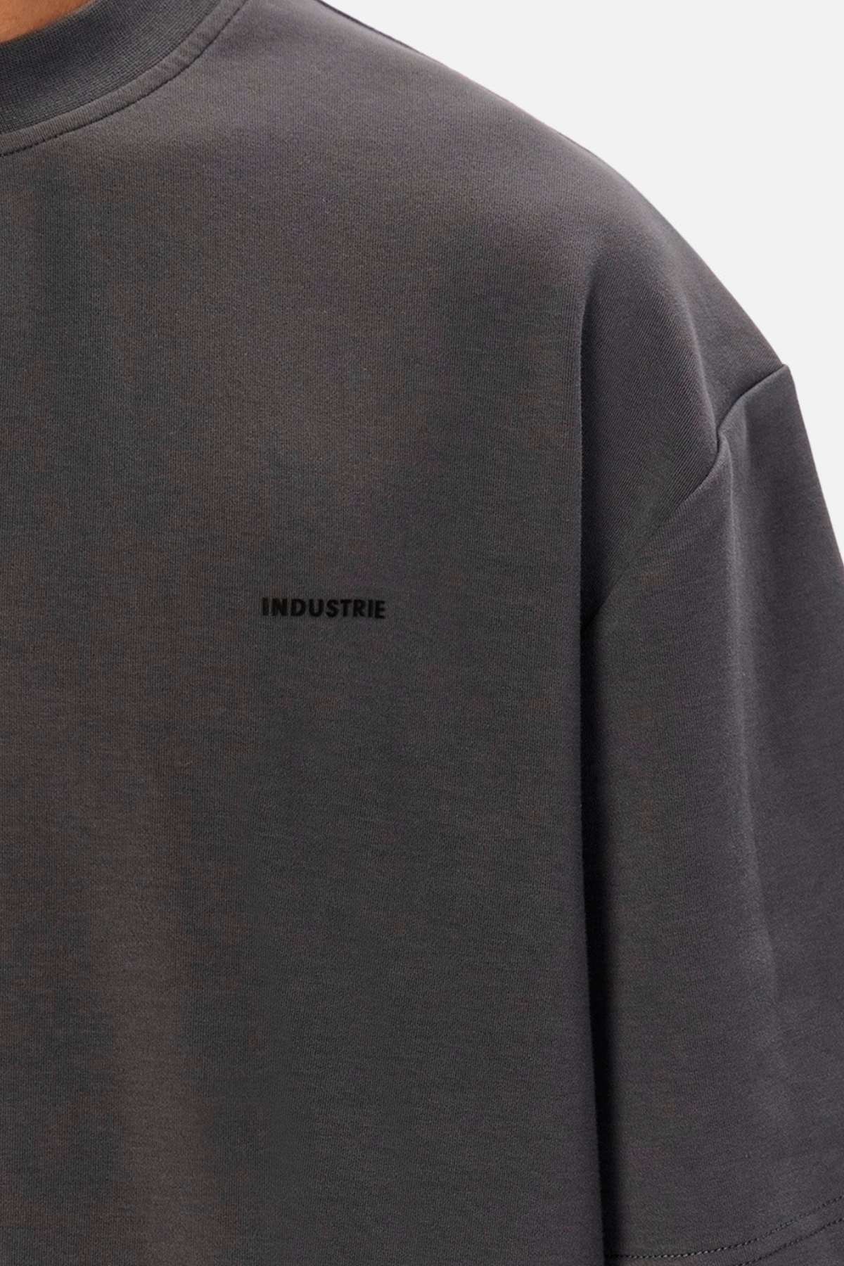 A close-up of a dark grey T-shirt, capturing details of the chest and sleeve, reveals the word "INDUSTRIE" printed in small, black capital letters on the left side of the chest. The fabric exhibits a soft and slightly textured appearance, typical of Industrie The Newport Tee made from 100% cotton.