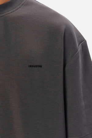 A close-up of a dark grey T-shirt, capturing details of the chest and sleeve, reveals the word "INDUSTRIE" printed in small, black capital letters on the left side of the chest. The fabric exhibits a soft and slightly textured appearance, typical of Industrie The Newport Tee made from 100% cotton.