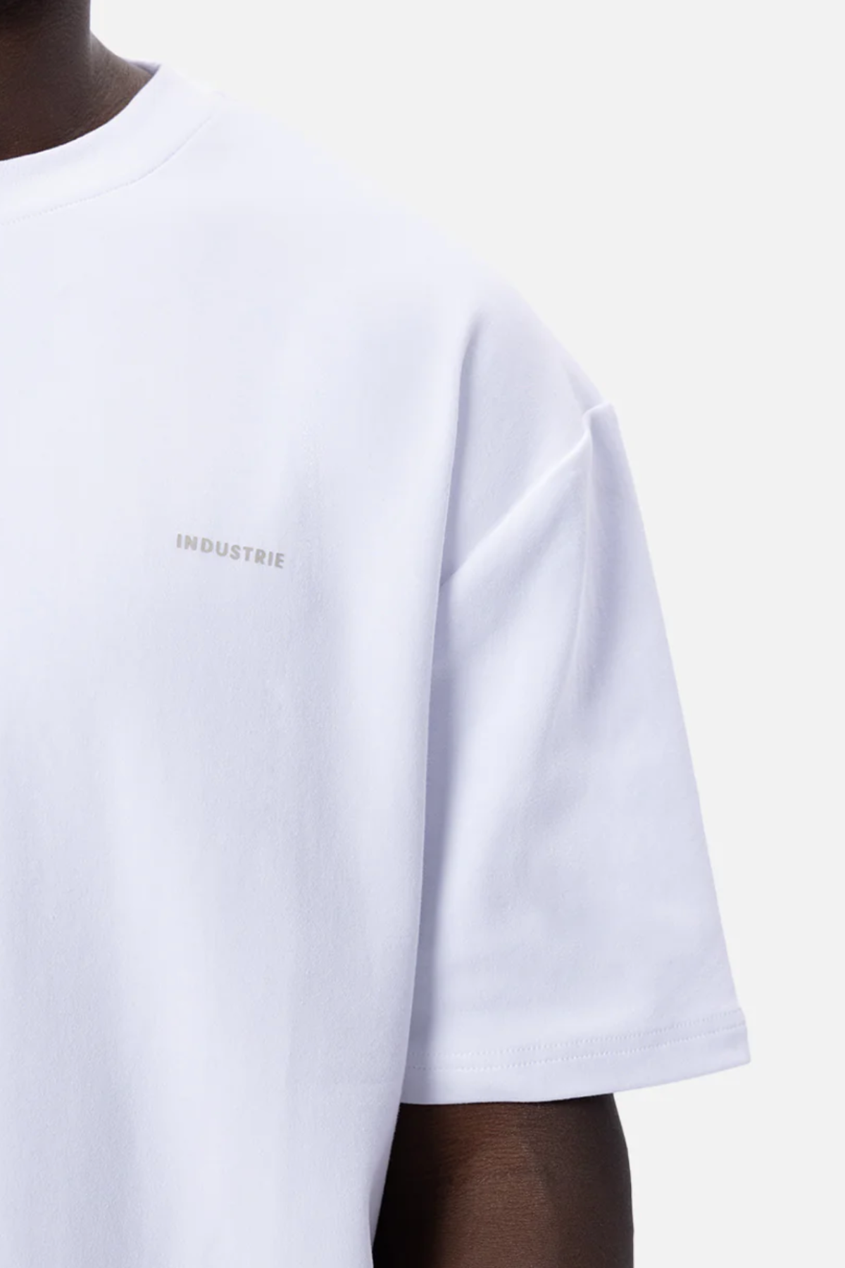 A close-up of a person wearing an oversized Industrie The Newport Tee, crafted from plain white 100% cotton with short sleeves. The t-shirt features the word "INDUSTRIE" subtly printed in small, light gray letters on the left side of the chest. The background is plain and white.