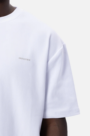 A close-up of a person wearing an oversized Industrie The Newport Tee, crafted from plain white 100% cotton with short sleeves. The t-shirt features the word "INDUSTRIE" subtly printed in small, light gray letters on the left side of the chest. The background is plain and white.
