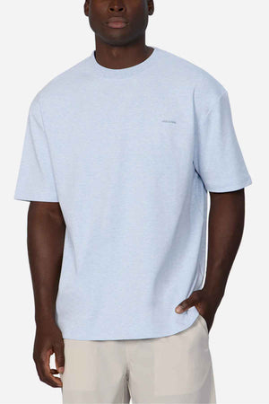 A person is wearing an oversized fit, light blue short-sleeve T-shirt and beige shorts, standing against a plain white background. The person has one hand in their pocket, and there is a small, subtle logo on the left side of the chest area of Industrie The Newport Tee.