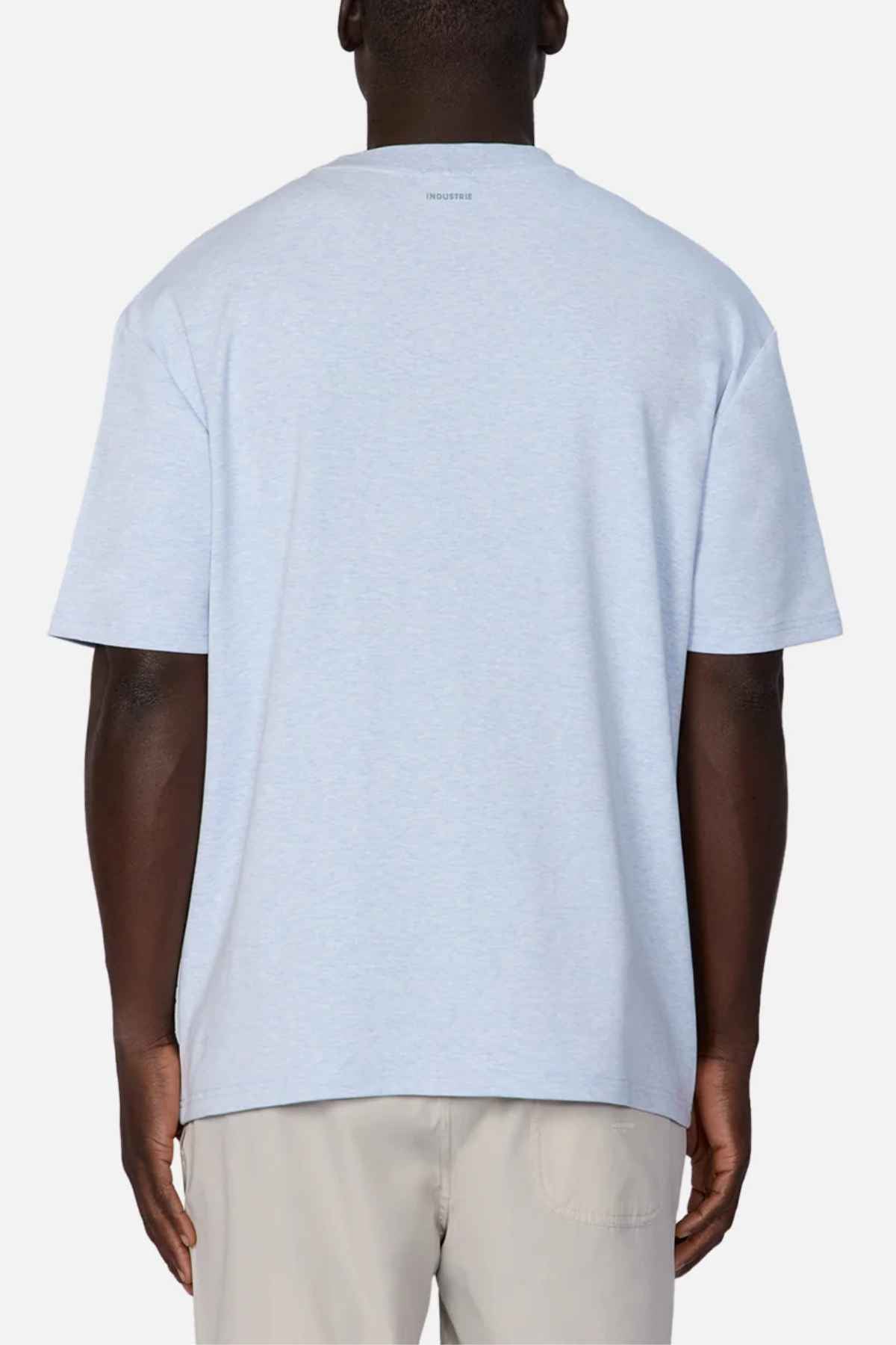 A man with dark skin is standing with his back to the camera, wearing a light blue short-sleeved T-shirt and beige pants. The T-shirt, named Industrie The Newport Tee, features a small, subtle logo in the upper center of the back near the neckline and fits him perfectly. The background is plain white.