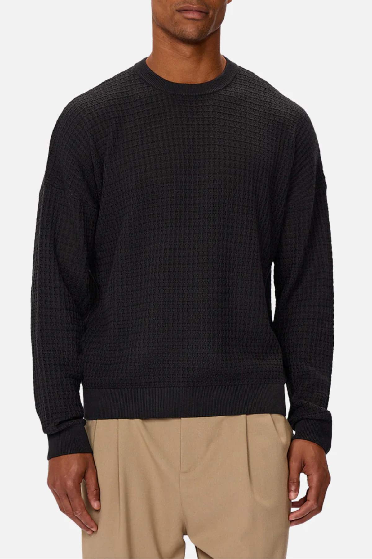 A man wears the Industrie The Nunez Long Sleeve Knit, a textured black crewneck sweater, paired with beige pleated pants, standing against a plain light gray background. His head is partially out of frame. This versatile knit suits any wardrobe.
