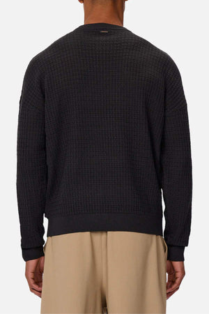 A person is shown from the back against a plain light background wearing the Industrie The Nunez Long Sleeve Knit, a textured black 100% cotton sweater, paired with beige pants.