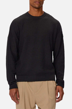 A man wears the Industrie The Nunez Long Sleeve Knit, a textured black crewneck sweater, paired with beige pleated pants, standing against a plain light gray background. His head is partially out of frame. This versatile knit suits any wardrobe.
