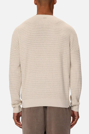 Back view of a person wearing Industrie The Nunez Long Sleeve Knit, a cream-colored, 100% cotton sweater with a small square pattern and a loose fit, paired with taupe pants against a plain white background.