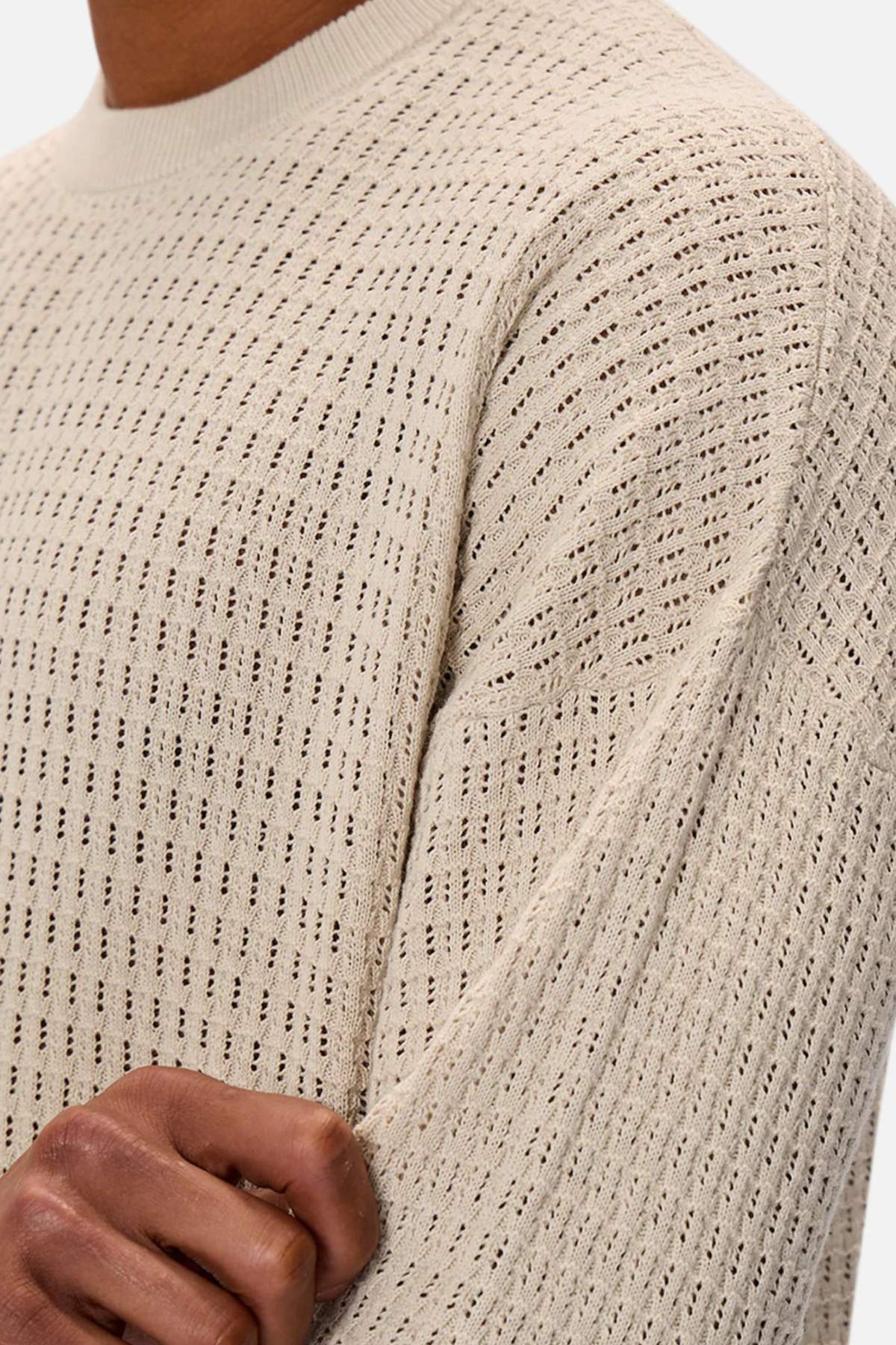 A person is wearing the Industrie The Nunez Long Sleeve Knit, a textured light beige sweater. Their hand gently holds the elbow, highlighting its open-weave pattern and 100% cotton composition.
