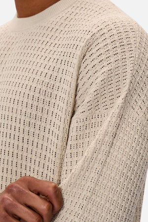 A person is wearing the Industrie The Nunez Long Sleeve Knit, a textured light beige sweater. Their hand gently holds the elbow, highlighting its open-weave pattern and 100% cotton composition.