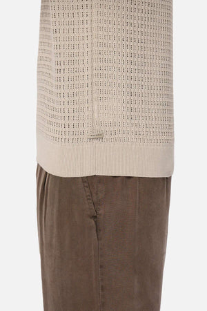 A close-up side view shows someone wearing a beige Industrie The Nunez Long Sleeve Knit sweater paired with brown corduroy pants, highlighting the intricate design and 100% cotton texture.