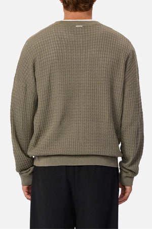 A person wears the Industrie The Nunez Long Sleeve Knit, an olive-green sweater with a woven pattern made of 100% cotton, featuring ribbed cuffs and hem. It's paired with black pants and set against a plain white background.