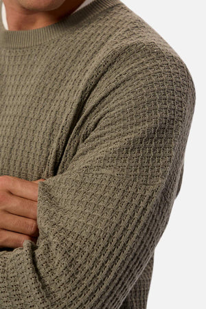 A person wears the Industrie The Nunez Long Sleeve Knit sweater in textured olive green, layered over a white shirt. Their arms are crossed, showcasing the grid-like pattern in 100% cotton with ribbed cuffs against a neutral background.