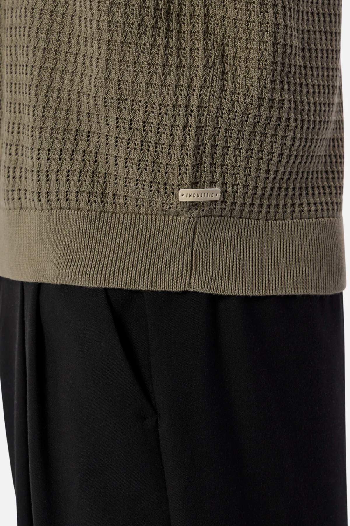 Close-up of the Industrie The Nunez Long Sleeve Knit, a green textured sweater with ribbed hem and cuffs, featuring a small metal tag engraved with "INDUSTRY." Made from 100% cotton, this long sleeve knit pairs perfectly with black pants.