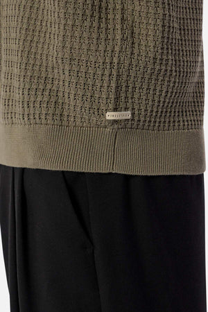 Close-up of the Industrie The Nunez Long Sleeve Knit, a green textured sweater with ribbed hem and cuffs, featuring a small metal tag engraved with "INDUSTRY." Made from 100% cotton, this long sleeve knit pairs perfectly with black pants.