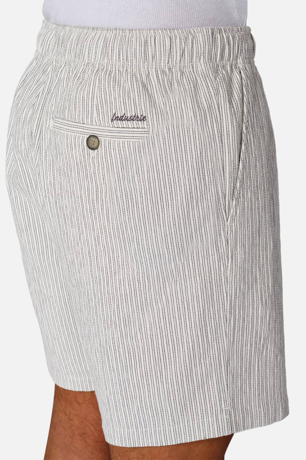 A person models Industrie The Ortenza Short—light gray and white vertically striped men's cotton shorts with an elastic waistband, side pocket, and buttoned back pocket—shown from the side against a plain background.