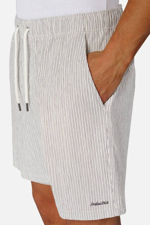 A person wears light-colored, vertically striped Industrie The Ortenza Short with a hand in the pocket, featuring a subtle embroidered logo near the hem, paired with a crisp white shirt.