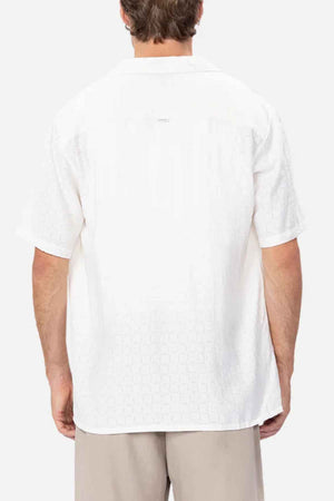 Dressed in an Industrie The Ortigia Short Sleeve Shirt crafted from breathable fabric with a subtle pattern on its white short-sleeve surface, the person stands back to the camera. Their look is effortlessly enhanced by beige pants, all set against a plain white background, embodying casual elegance.