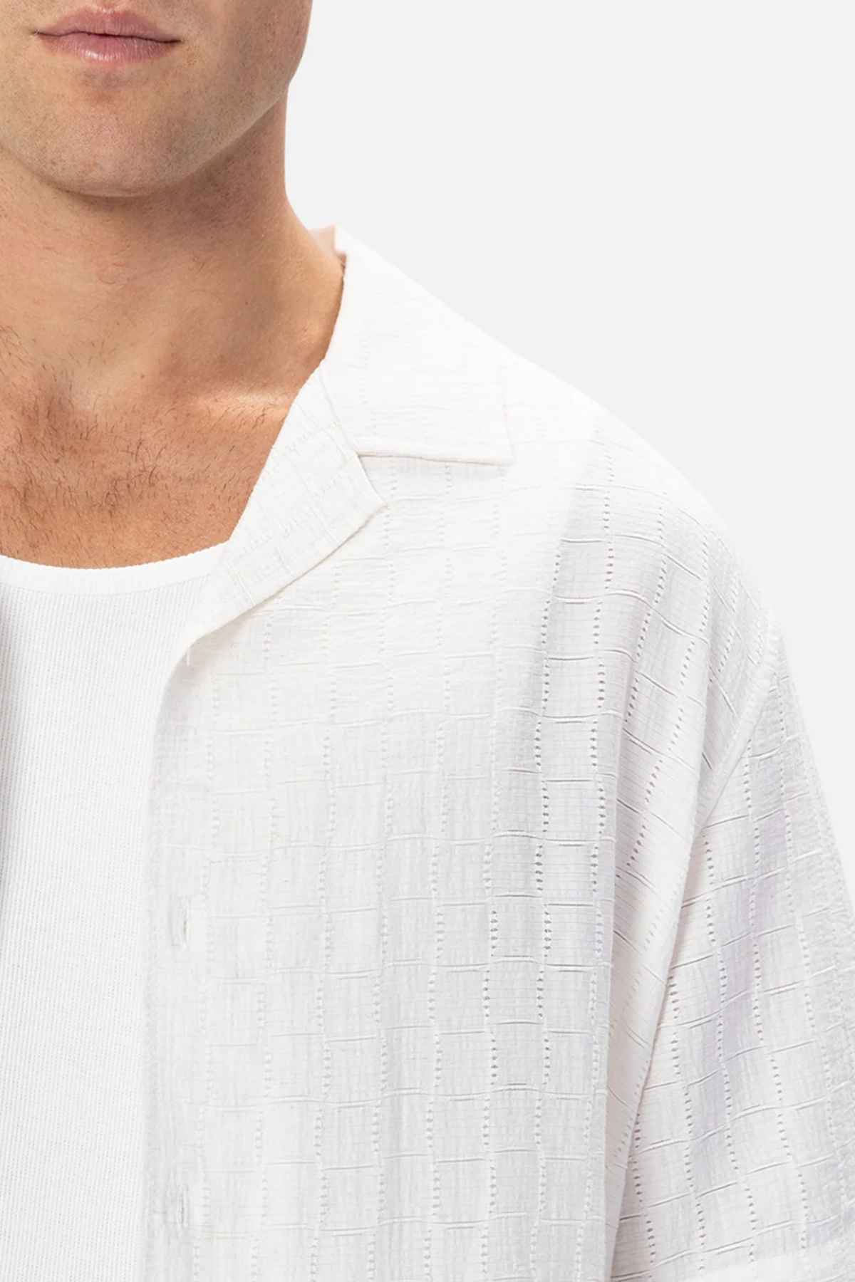 A person wears a textured white button-up, such as the Industrie The Ortigia Short Sleeve Shirt, over a white ribbed tank top. The focus is on their chest and shoulder area against a plain, light background.
