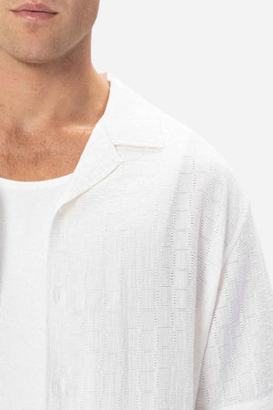 A person wears a textured white button-up, such as the Industrie The Ortigia Short Sleeve Shirt, over a white ribbed tank top. The focus is on their chest and shoulder area against a plain, light background.