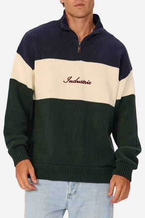 A person is wearing the Industrie The Osbourne Zip Neck Knit, a timeless piece featuring wide navy, cream, and dark green horizontal stripes. The cream stripe boasts the word "Industrie" embroidered in red cursive. Paired with light blue jeans, this outfit is both stylish and comfortable in 100% cotton.