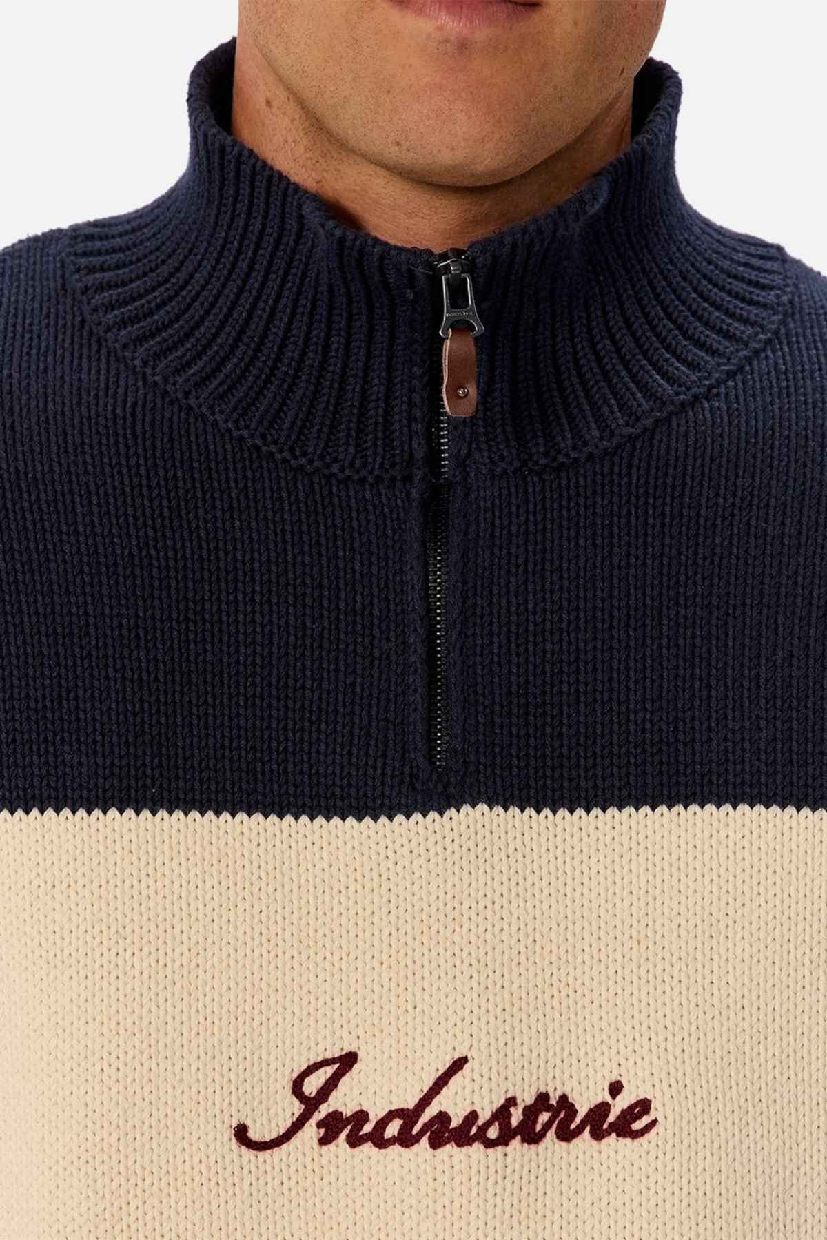 A person wearing the Industrie The Osbourne Zip Neck Knit, a classic sweater crafted from 100% cotton. This half-zip knit showcases a ribbed collar, with its upper section in dark blue and the lower section in beige, while "Industrie" is elegantly embroidered in red on the chest. The person's lower face is partially visible.