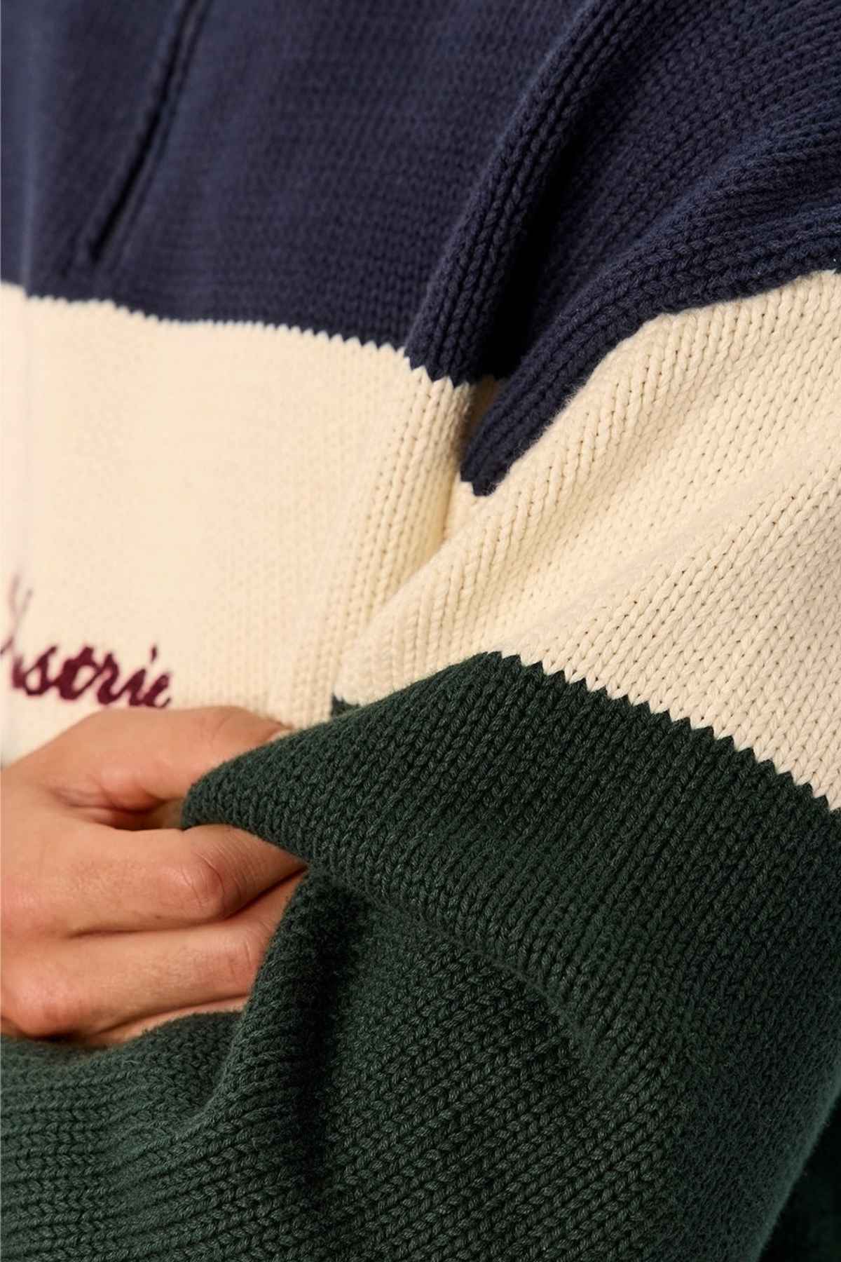 Close-up of a person wearing the Industrie The Osbourne Zip Neck Knit, a timeless knitted sweater featuring three color blocks: dark blue at the top, cream in the middle, and forest green at the bottom. The individual gently holds the green sleeve, showcasing this 100% cotton garment. Only a section of their torso and arm is visible.