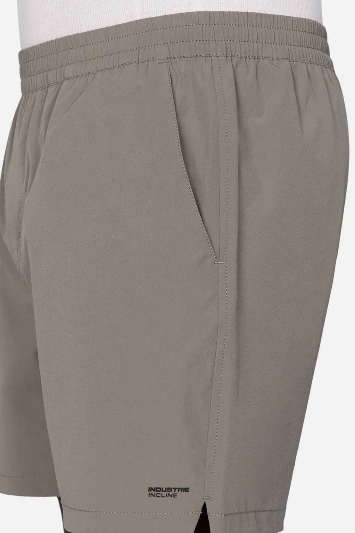 A close-up of a person wearing "Industrie The Owens Short" in light gray, featuring an elasticized waistband and the branding "Industrie Incline" near the hem. These above-knee-length shorts boast quick-drying fabric and have a visible side pocket. The person is sporting a white shirt that is slightly tucked in.
