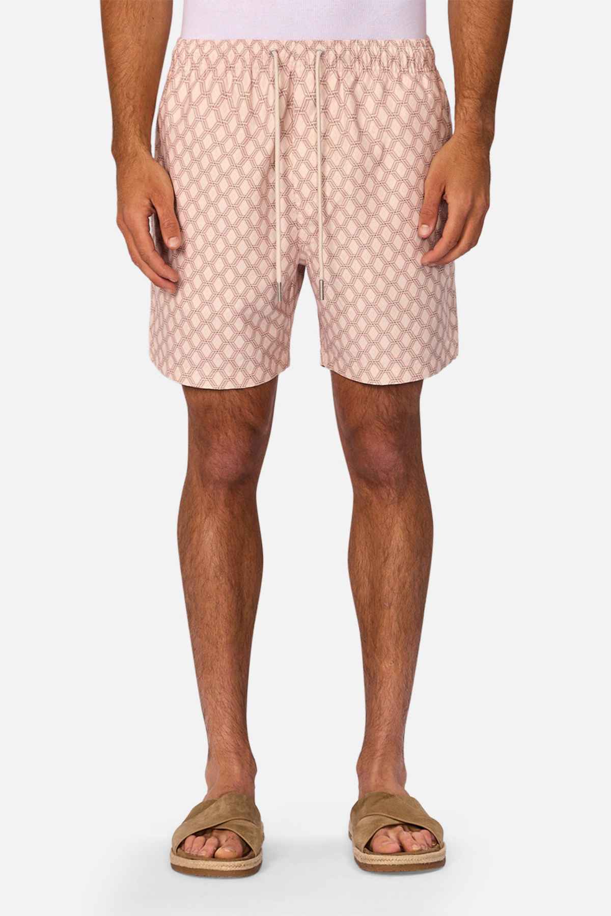 A man, visible from shoulders to feet, wears a white shirt, tan slip-on sandals, and Industrie The Panama Swim Short in light pink pattern, standing against a plain white background to highlight stylish men's swimwear.